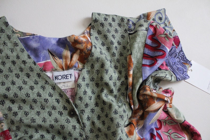 May include: A multi-colored, patterned dress with a V-neckline and short sleeves. The dress has a green, floral, and paisley print. The label on the dress reads "KORET".