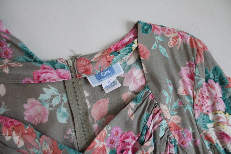 Op de afbeelding: Vintage jurk met een bloemenpatroon in roze, rood en teal op een grijze achtergrond. De jurk heeft een V-hals en een ritssluiting. Het label leest "Qps" en maat 12.