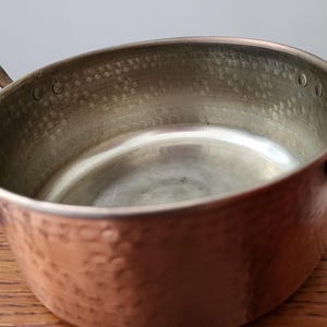 May include: A copper cooking pot with two brass handles. The pot has a hammered texture on the exterior and a smooth, reflective interior. The pot is sitting on a wooden surface.