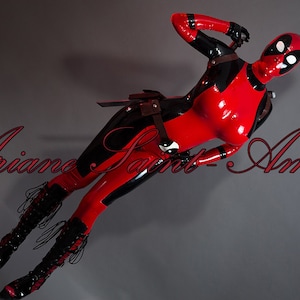 Può includere: Una donna in un completo di lattice rosso e nero con una maschera di Deadpool, posa con una spada. Il testo "Ariane Saint-Amour" è visibile sullo sfondo.