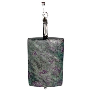 May include: A rectangular pendant with a silver bail and a natural stone body. The stone is primarily gray-green with small flecks of purple. The pendant has a polished finish and a simple, elegant design.