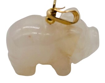 May include: A light-colored, translucent pig pendant with a gold-colored bail. The pig is carved with rounded features and a smooth surface. The bail is attached to the top of the pig's head.
