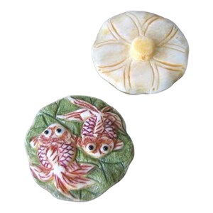 Op de afbeelding: Twee decoratieve keramische stukken. Eén is een crèmekleurig, bloemvormig deksel. De andere is een groen, bladvormig schaaltje met twee oranje en witte goudvissen. De goudvissen hebben gedetailleerde schubben en vinnen.