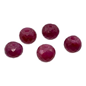 May include: Five faceted, round ruby beads. Each bead is a deep red color with a hole drilled through the center. The beads have a polished, reflective surface, and are arranged against a white background.