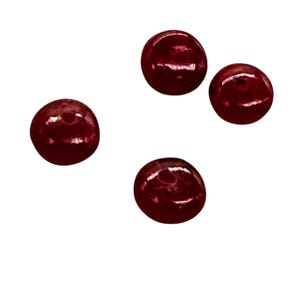 May include: Four round, dark red beads with a glossy finish are scattered against a white background. The beads appear to be made of a smooth, reflective material, catching the light and creating highlights.
