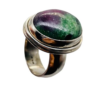 May include: A silver ring featuring a large, oval gemstone. The gemstone displays a mix of colors, including deep purple, vibrant green, and hints of white. The ring has a thick band and a bezel setting that securely holds the stone.