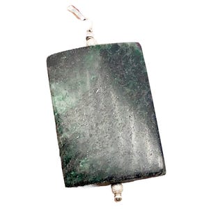 May include: A rectangular pendant with a dark green and gray mottled appearance. The pendant has a silver-colored bail at the top and a silver-colored bead at the bottom. The stone has a textured surface and is rectangular in shape.