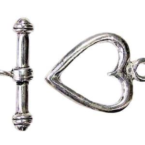 May include: A silver-toned toggle clasp set. The clasp features a bar and a heart-shaped loop. The heart has a decorative design. The clasp is likely for jewelry making or crafts.