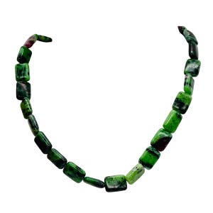 May include: A necklace composed of rectangular green and dark green beads. The beads are strung together to form a necklace. The necklace is displayed against a white background. The beads have a polished, glossy finish.