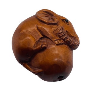 May include: A detailed wooden carving of a curled-up elephant. The elephant is brown with visible wood grain and a small, dark eye. The trunk is curled, and the ears are visible. The carving is smooth and rounded, with a hole at the bottom.