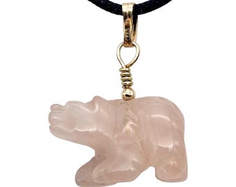 May include: A carved rose quartz bear pendant suspended from a gold-toned metal bail and a black cord. The bear is depicted in a walking stance, showcasing the translucent, pale pink stone with smooth, polished surfaces and defined features.