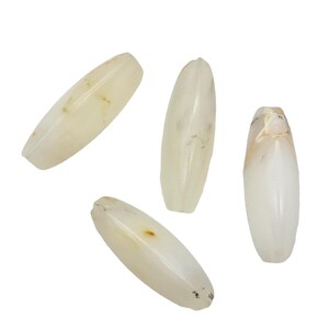 May include: Four oblong, off-white beads with a polished, smooth surface. Each bead has a slightly irregular shape and subtle variations in color, with hints of tan and brown. The beads are arranged against a plain white background.