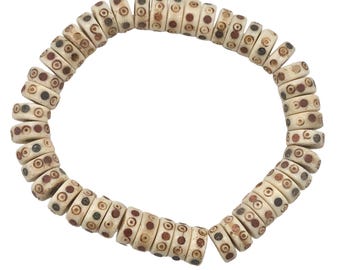 May include: A cream-coloured bracelet made of cylindrical beads. Each bead has small, dark brown, red, and black circular patterns. The bracelet is arranged in a circular shape against a white background.