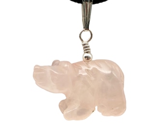 May include: A rose quartz bear pendant hangs from a black cord. The bear is carved with detail, showcasing its form. The pendant is attached to a silver-colored bail. The background is white.
