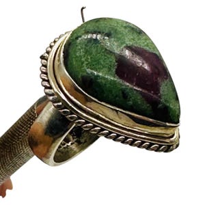 Puede incluir: Un anillo con una piedra en forma de pera, de color verde moteado y púrpura oscuro, engastada en una banda de metal plateado. El engaste incluye un borde decorativo de cuerda trenzada que rodea la base de la piedra, montada sobre un aro metálico liso.