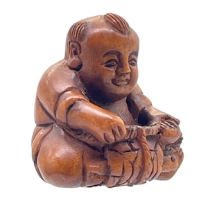 May include: A carved wooden figurine of a seated figure with a smiling expression. The figure is depicted with a rounded body and is holding a basket or container. The wood has a warm, brown tone, and the carving details are visible.