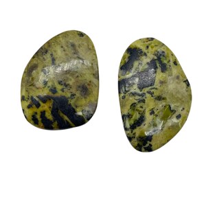 May include: Two polished, freeform gemstones. The stones have a mottled appearance with a base color of olive green, accented by black and yellow patterns. The gemstones are set against a white background.