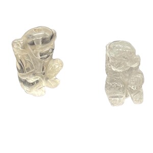 May include: Two clear quartz crystal carvings of small, seated animal figures. The carvings are detailed, with visible features and a smooth, polished appearance. The figures are positioned against a white background.