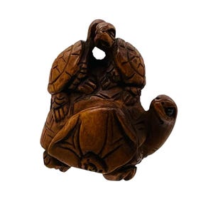 Op de afbeelding: Een gedetailleerde houten sculptuur van drie schildpadden. De schildpadden zijn op elkaar gestapeld, waarbij de bovenste twee elkaar lijken te omarmen. De sculptuur heeft een warme bruine kleur en heeft ingewikkelde details.