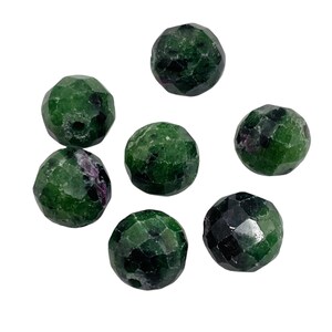 May include: Seven faceted, round gemstones. The stones are a deep green with flecks of a darker color. The gemstones are scattered on a white background. The gemstones are likely used for jewelry making or other crafts.