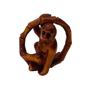 May include: A carved wooden monkey figurine, brown in color, holding a bamboo ring. The monkey has detailed facial features and is posed in a playful position. The bamboo ring is segmented, adding to the intricate design of the sculpture.