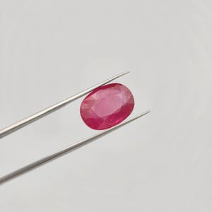 May include: A vibrant, oval-cut ruby gemstone held by silver tweezers. The ruby displays a rich, deep red hue with subtle internal patterns. The tweezers are positioned against a plain, light background, highlighting the gem's color and shape.