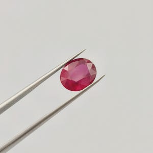 May include: A faceted, oval-shaped ruby gemstone, held by silver tweezers. The ruby displays a deep red hue with internal light reflections. The background is a plain, light gray.