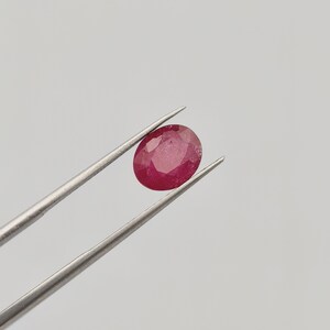 May include: A vibrant, oval-cut ruby gemstone, held by silver tweezers against a plain white background. The ruby displays a deep red hue with internal inclusions, showcasing its natural characteristics. This gemstone is a focal point for jewelry design.