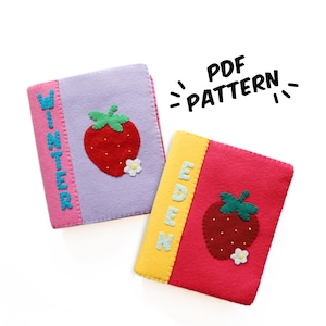 Puede incluir: Dos cubiertas de libros de fieltro. Una es lavanda con lomo rosa y el nombre "WINTER" en azul. La otra es roja con lomo amarillo y el nombre "EDEN" en azul. Ambas tienen una aplicación de fresa.
