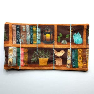 May include: A rectangular fabric item featuring a printed design of a bookshelf. The design includes various books, a bird, a potted plant, and a glowing lantern. The color palette consists of browns, greens, and blues.