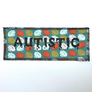 以下が含まれることがあります： 白とオレンジの蛙の模様がついたターコイズブルーの布のパッチ。パッチの中央に黒い文字で「AUTISTIC」と印刷されています。