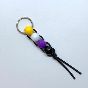 Puede incluir: Un llavero con un cordón negro y cuentas de colores amarillo, blanco, morado y negro.