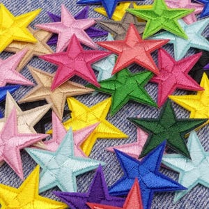 May include: A selection of embroidered star patches in a variety of colours, such as yellow, pink, green, blue, and purple. The patches are arranged on a denim fabric background, creating a vibrant and textured display. Each star has a detailed stitched outline.