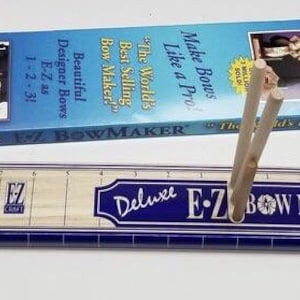 May include: A wooden bow making tool with a blue and white label that reads "Deluxe E-Z Bow Maker". The tool has a ruler and a pool holder. The tool is on a white surface.