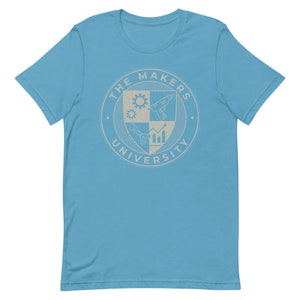 May include: A light blue t-shirt with a white graphic of a crest. The crest has a shield with four sections. Each section has a different symbol: a gear, a drill, scissors, and a graph. The crest is surrounded by a circle with the text "THE MAKERS UNIVERSITY".