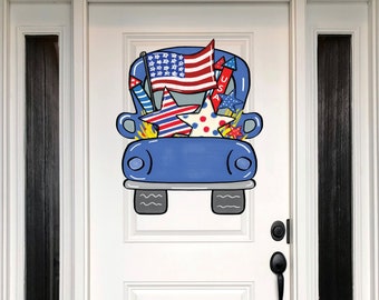May include: A blue truck-shaped wooden door hanger decorated with patriotic symbols. The truck bed is filled with a waving American flag, fireworks, and star-shaped decorations in red, white, and blue. The door hanger is displayed on a white door.