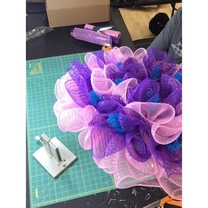 May include: A pink and purple mesh wreath with blue accents is being made on a green cutting mat. The wreath is being made with a wreath making tool. There are also scissors and other tools on the mat.