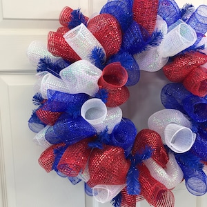 Puede incluir: Una corona de malla roja, blanca y azul con detalles de brillo. La corona está hecha de múltiples capas de malla y está decorada con plumas azules y blancas.