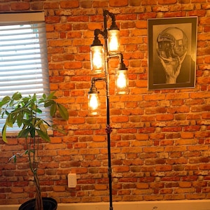 May include: A floor lamp made of industrial pipes with four light bulbs in glass jars. The lamp is standing on a wooden floor in front of a brick wall.