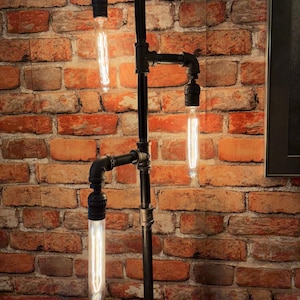 May include: A black industrial-style floor lamp with three exposed Edison-style light bulbs. The lamp is constructed from black pipes and fittings, and is mounted against a red brick wall. The bulbs are long and cylindrical.