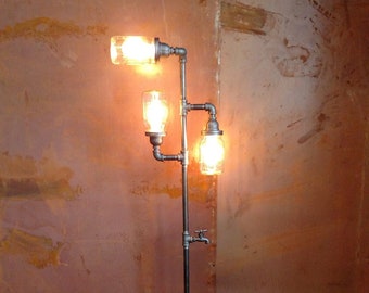 Könnte beinhalten: Eine Stehlampe aus schwarzen Metallrohren mit drei Glasbehältern als Leuchten. Die Lampe ist eingeschaltet und die Behälter leuchten. Die Lampe steht auf einem Holzboden vor einer rostigen Metallwand.