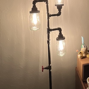 May include: A floor lamp made from black pipes with four mason jar light fixtures. The lamp has a red valve on the side and is lit with warm white light bulbs.