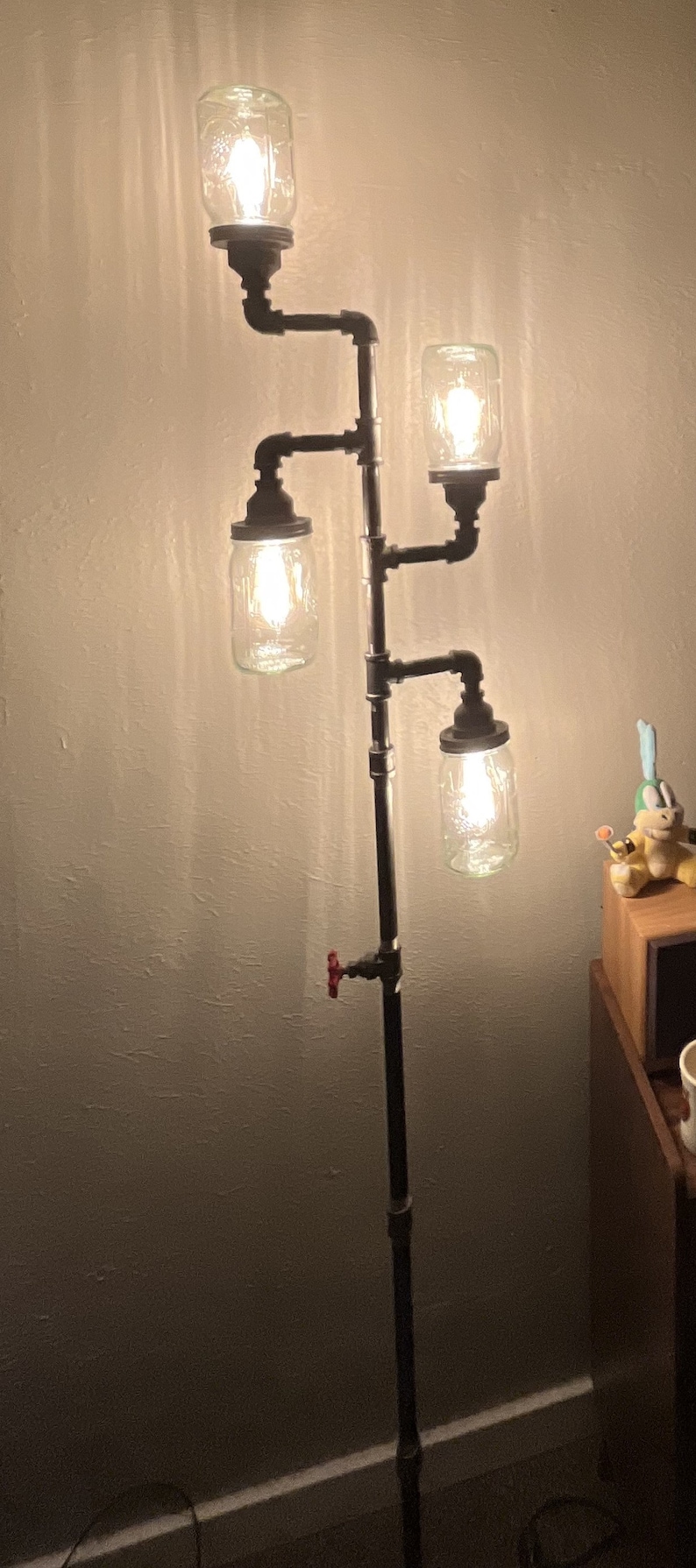May include: A floor lamp made from black pipes with four mason jar light fixtures. The lamp has a red valve on the side and is lit with warm white light bulbs.