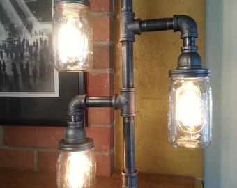 May include: A unique, industrial-style floor lamp constructed from black metal pipes. Three clear glass mason jars serve as light fixtures, each housing a glowing bulb. The lamp stands against a brick wall and wooden surface, creating a rustic aesthetic.