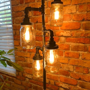 May include: A four-light industrial style floor lamp with exposed black pipes and four clear glass mason jars with light bulbs. The lamp is set against a brick wall.