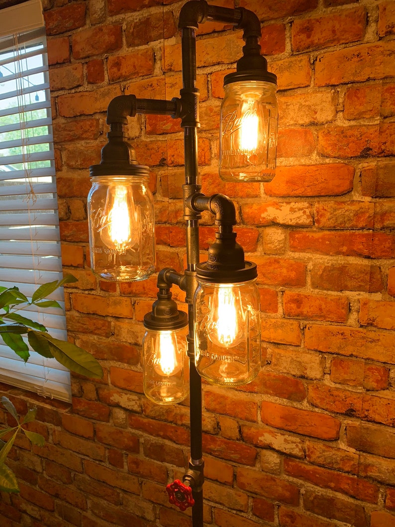 May include: A four-light industrial style floor lamp with exposed black pipes and four clear glass mason jars with light bulbs. The lamp is set against a brick wall.