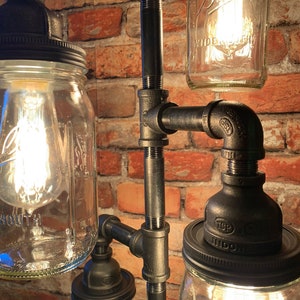May include: A black metal pipe lamp with four mason jars, each with a light bulb inside. The jars are attached to the pipes with black metal caps. The lamp is set against a brick wall.