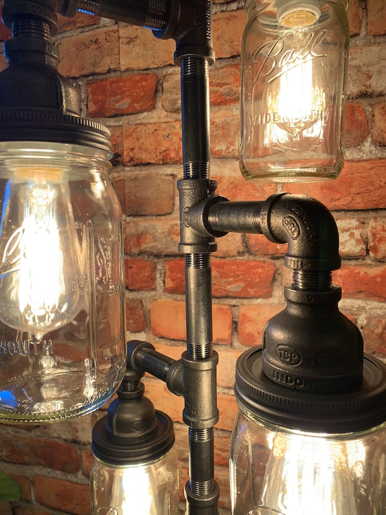 May include: A black metal pipe lamp with four mason jars, each with a light bulb inside. The jars are attached to the pipes with black metal caps. The lamp is set against a brick wall.