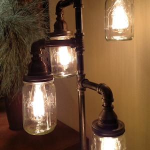 May include: A floor lamp made from industrial pipes and mason jars. The lamp has four lights, each with a clear glass jar and a light bulb. The lamp is on a wooden surface.