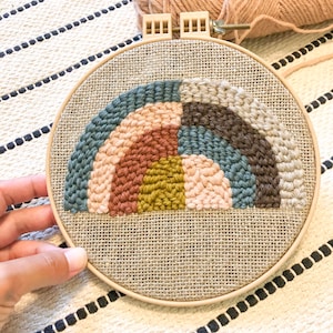 May include: A rainbow-colored yarn embroidery hoop art project with a variety of colors including brown, pink, blue, green, and yellow. The hoop is on a striped fabric surface.